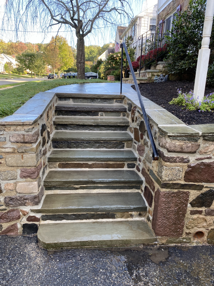 Stone masonry steps with a secure handrail leading up to a landscaped pathway, set in front of a house in a residential neighborhood.
